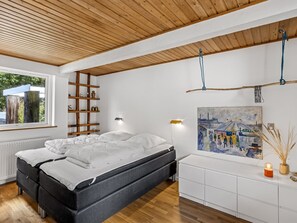 A modern bedroom with a wooden ceiling, a bed with white bedding, a bedside table, a wall-mounted shelf, and a painting.