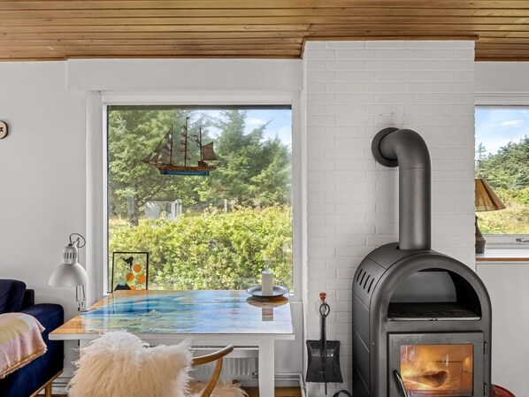 A cozy room with a wood-burning stove, a table with a blue tablecloth, and a view of greenery outside.