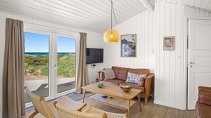 Una habitación con piso de madera, un sofá de cuero marrón, una mesa de centro de madera, sillas de mimbre, un televisor y una puerta que da a un balcón con vistas al mar.