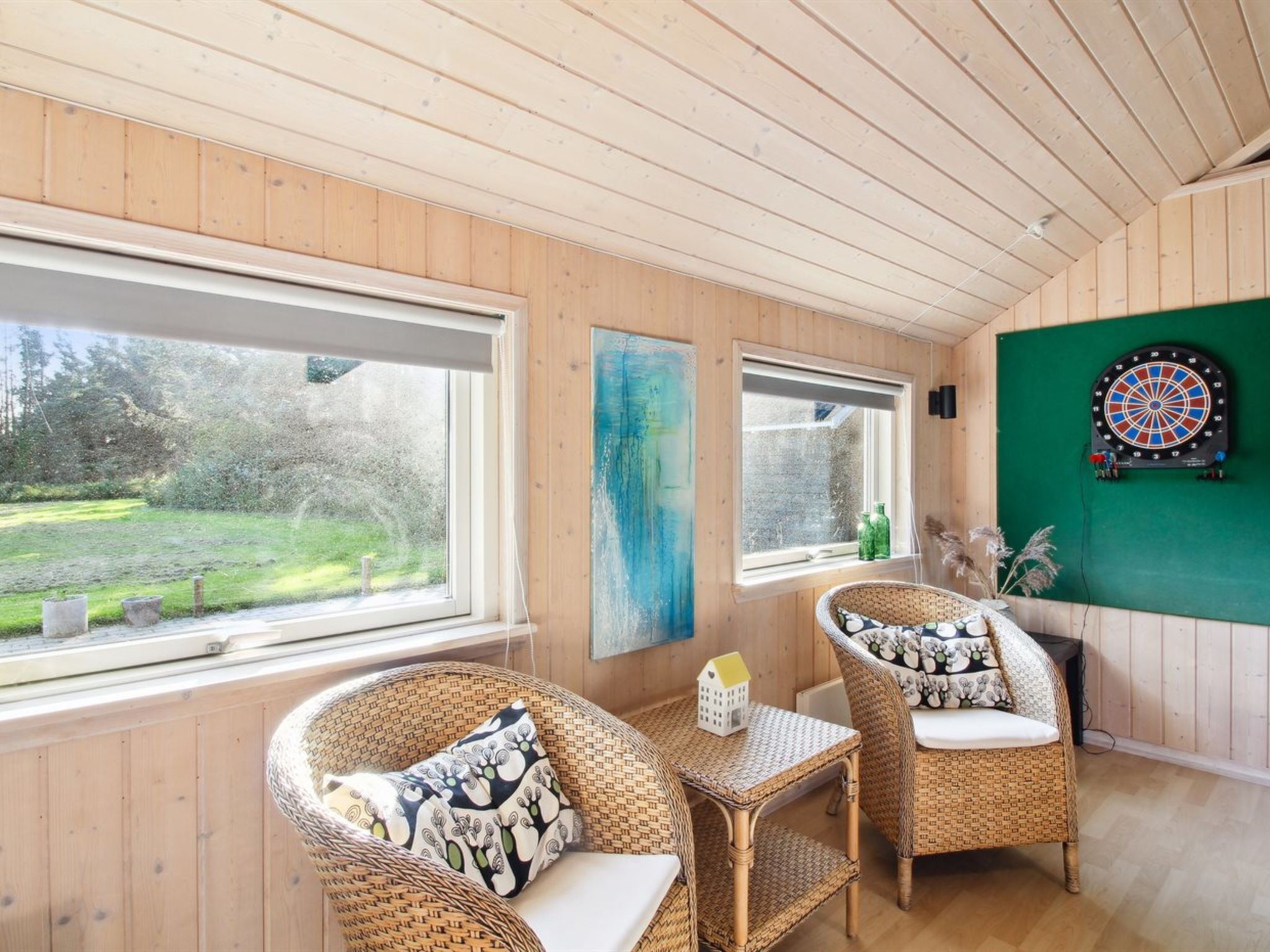 A cozy room with wooden walls and ceiling, featuring two wicker chairs with cushions, a small table, and a dartboard on the wall.