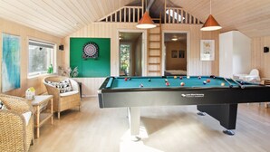 Une table de billard dans une pièce aux murs et au plafond en bois, un jeu de fléchettes et un fauteuil en osier.