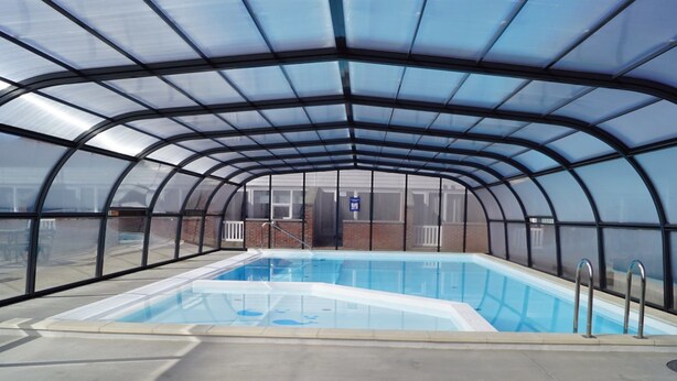 Una piscina techada con techo de vidrio, rodeada por un muro de ladrillo y una cerca metálica.