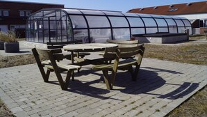 Estructura de invernadero con techo transparente, rodeada por un patio de ladrillo con bancos y mesas de madera.