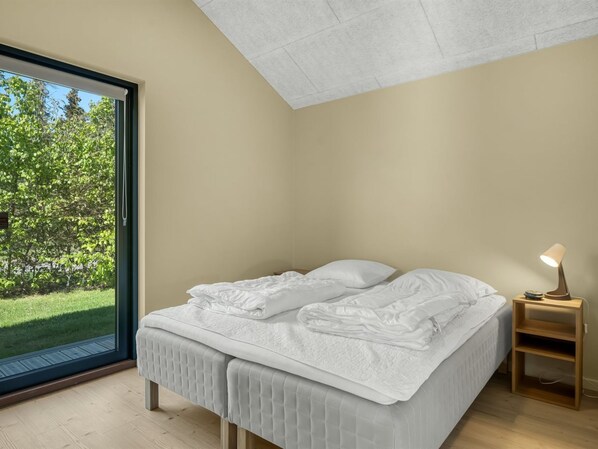 A bedroom with a large window, a bed with white linens, a bedside table with a lamp, and a view of greenery outside.