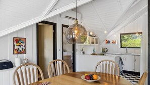 A dining area with a wooden table, chairs, and a fruit bowl. There is a kitchen visible in the background with white cabinets and a window.