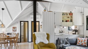 A modern living room with a high ceiling, a dining area with wooden chairs, a mustard-colored armchair with a white fur throw, a gray sofa with orange and black patterned pillows, and a small white table with a magazine and candle.