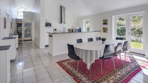 Une cuisine spacieuse dotée d’un îlot central, un coin repas avec une table ronde et des chaises, et une vue sur l’extérieur grâce à de grandes fenêtres.