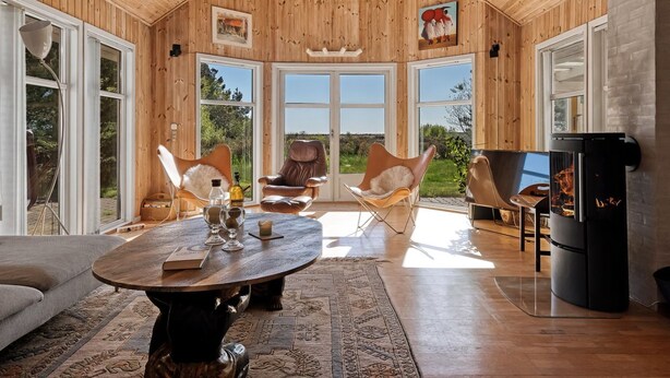 Une pièce spacieuse aux murs en bois et aux grandes fenêtres, avec une table ronde entourée de chaises, un canapé et une cheminée.