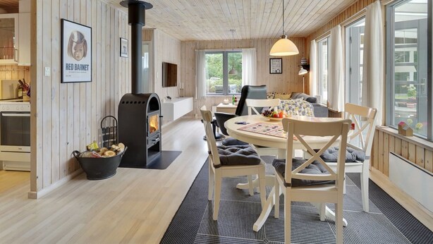 Un intérieur de chalet moderne avec un plafond lambrissé, une cheminée centrale, un coin repas avec une table et des chaises, et de grandes fenêtres laissant entrer la lumière naturelle.