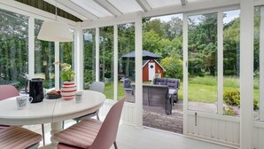 Une véranda dotée d’une table ronde, de chaises et d’un coin café. De grandes fenêtres offrent une vue sur un jardin et une grange rouge.