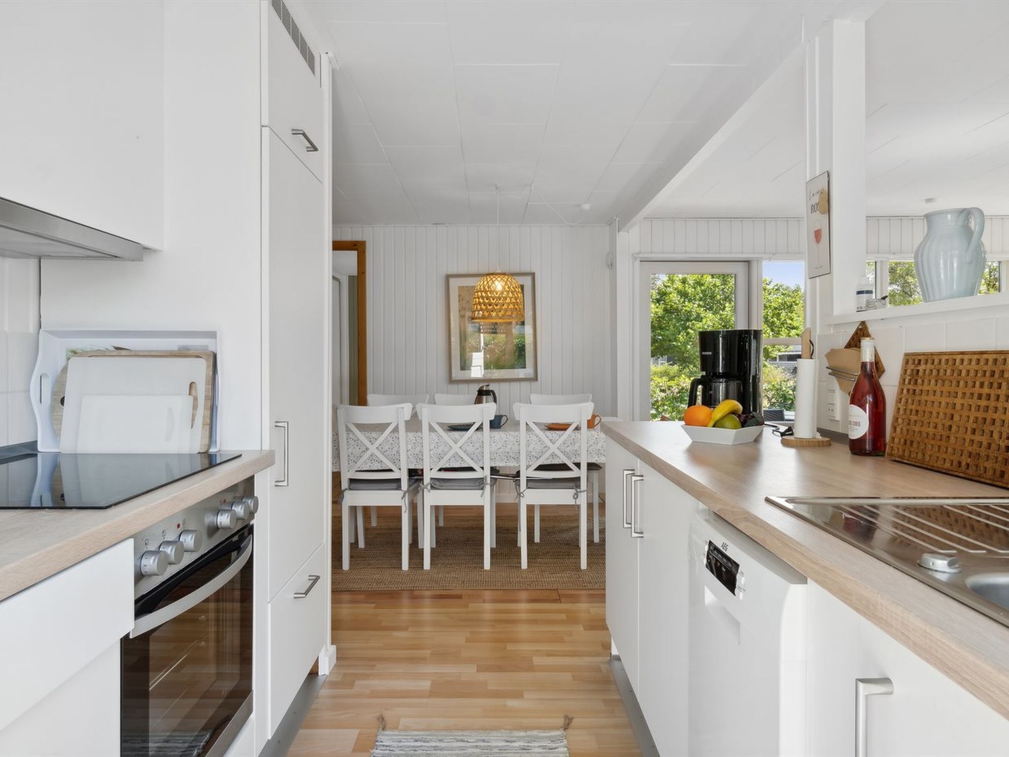 A modern kitchen with white cabinetry, a wooden countertop, and a built-in oven. There is a dining area with white chairs and a table, and a window providing a view of greenery outside.