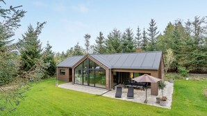 Ein modernes Haus mit großer Glasfront, einer Terrasse mit Gartenmöbeln und einem umgebenden Wald.