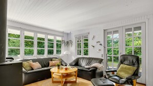 A modern living room with a sofa, a round table, a chair, and large windows offering a view of greenery.