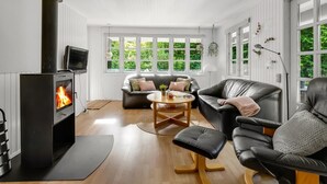 A modern living room with a wood-burning stove, leather furniture, and large windows offering a view of greenery.
