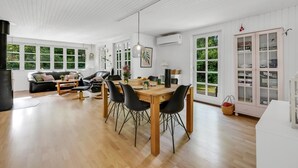 A spacious living area with a dining table, black chairs, a sofa, and a large window providing a view of greenery.
