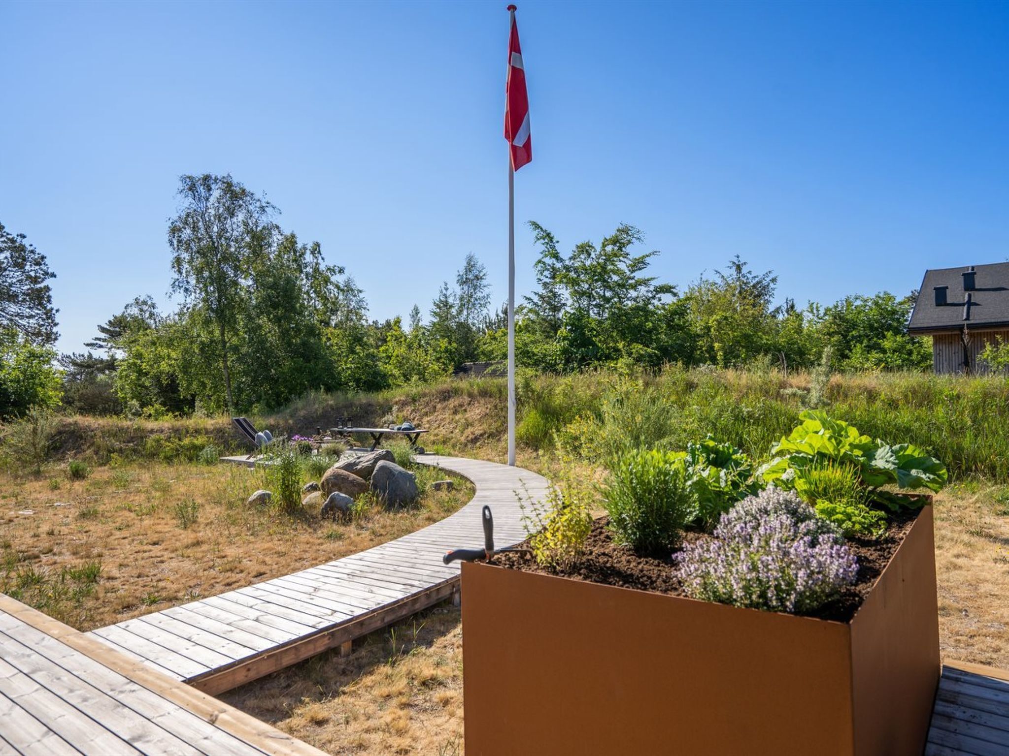 A wooden deck leading to a garden area with a flagpole and a building in the background.