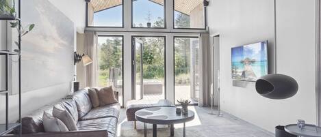 A spacious living area with a high ceiling, large windows, a modern sofa, a round coffee table, and a flat-screen TV.