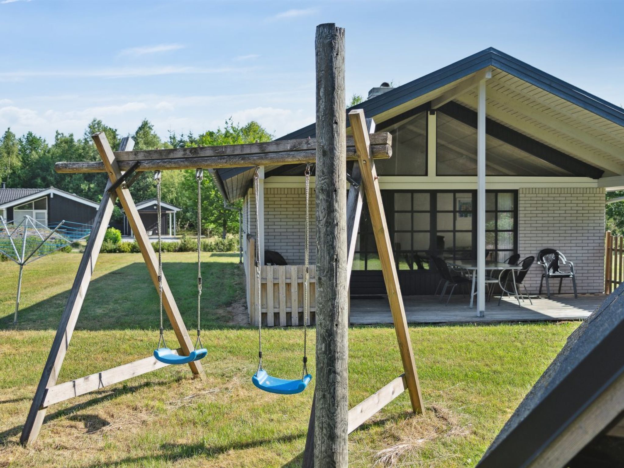 Un ensemble de balançoires en bois installé dans un jardin, avec un patio couvert et une maison en briques en arrière-plan.