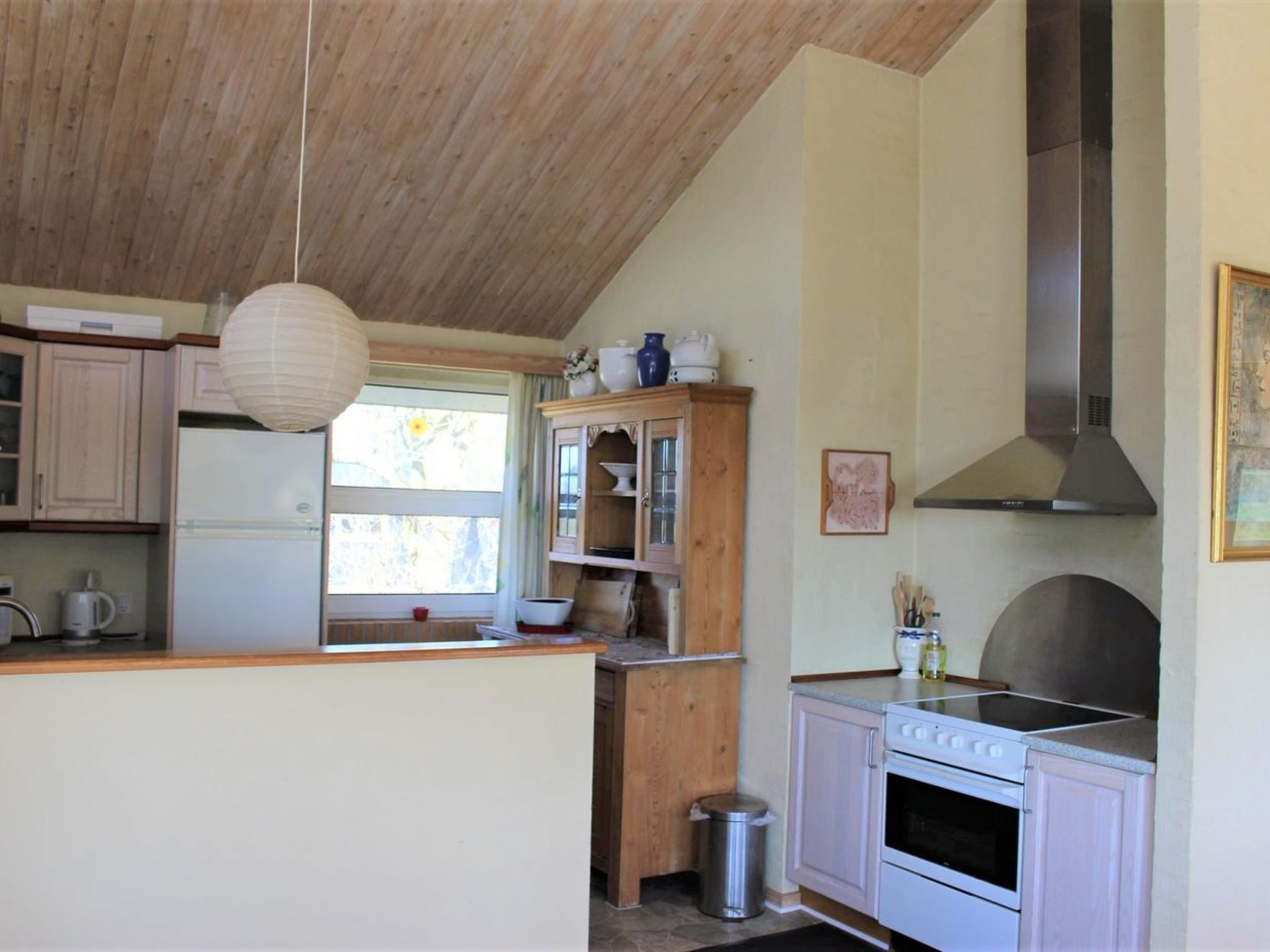 Une cuisine avec un plafond en bois, un réfrigérateur blanc, une cuisinière et une fenêtre donnant sur un paysage verdoyant.