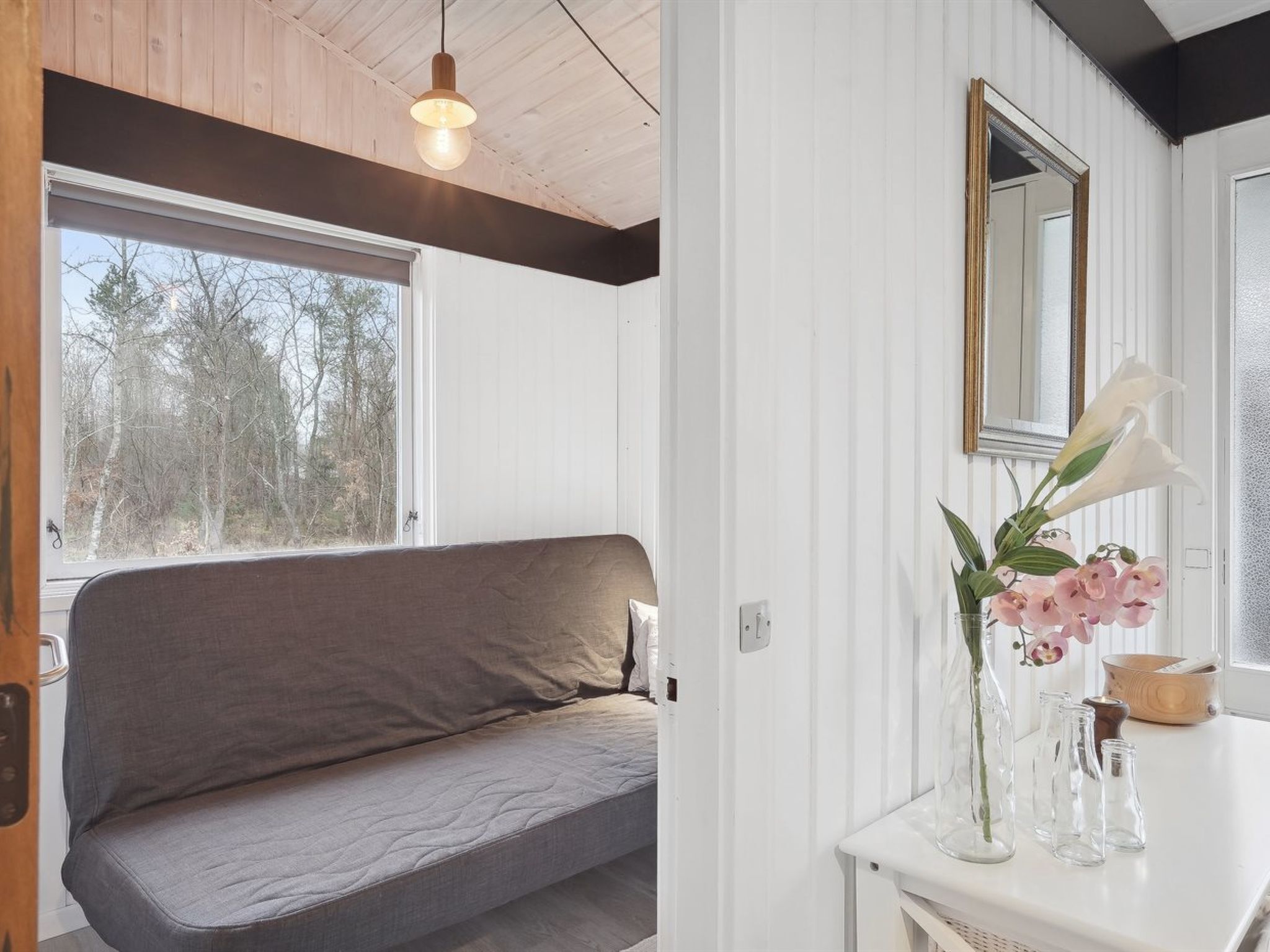 Ein Zimmer mit grauem Sofa, Holzboden, großem Fenster, weißer Wand, Spiegel und Blumen in einer Vase.