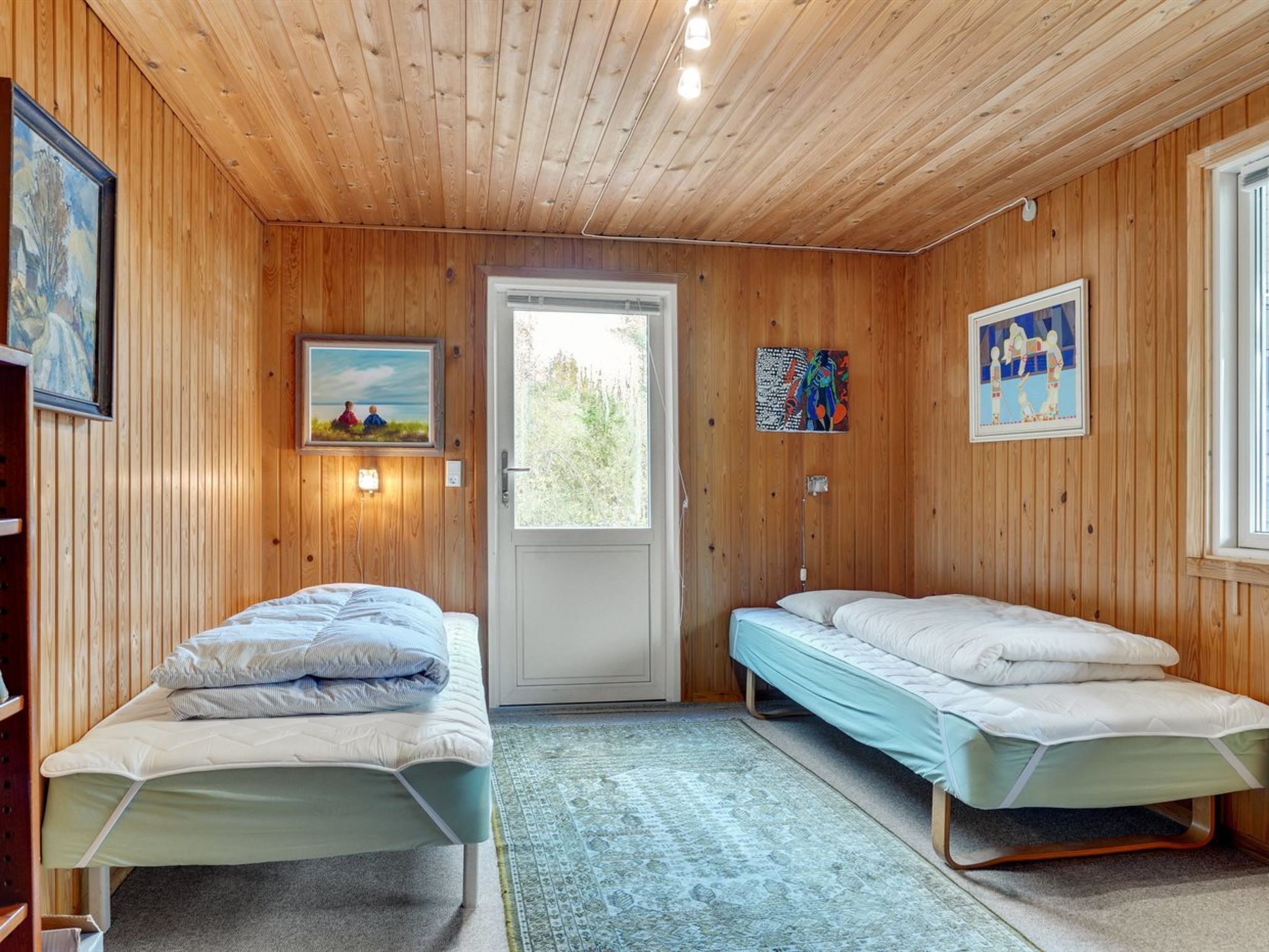 A room with two single beds, a wooden ceiling, a door leading outside, and artwork on the walls.