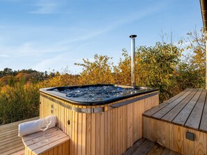 Un bain à remous en bois, doté d’une cheminée en métal, entouré d’une terrasse et de bancs en bois, le tout se fondant dans un décor automnal aux couleurs flamboyantes.