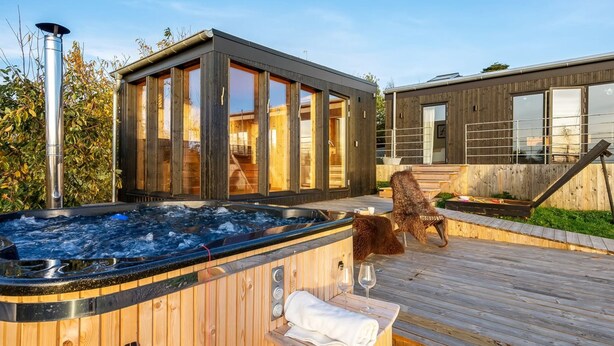 Et moderne udendørs område med jacuzzi, træterrasse, en træsauna og en tilbygning med glas.