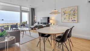 Un salon moderne avec un coin repas, de grandes fenêtres donnant sur la mer et une œuvre d’art contemporain accrochée au mur.