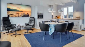 Una cocina moderna con armarios blancos, una zona de comedor con mantel azul y un cuadro grande de un atardecer sobre el agua.