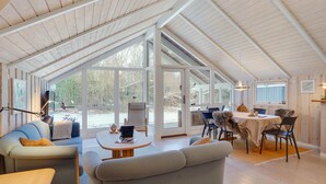 Un espacio amplio y bien iluminado, con techos altos y ventanas grandes, que incluye una zona de estar acogedora con sofá y sillones, una mesa de comedor y vistas al exterior.