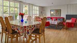Spiseområde med et rundt bord, stole og en blomstervase. En sofa og et billede ses i baggrunden.