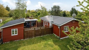 En rød træhytte med en indhegnet terrasse, en lille tilbygget garage og en parabantenne.