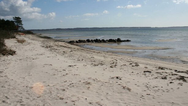 En sandstrand med en stenmole, klare himmel og land i det fjerne.