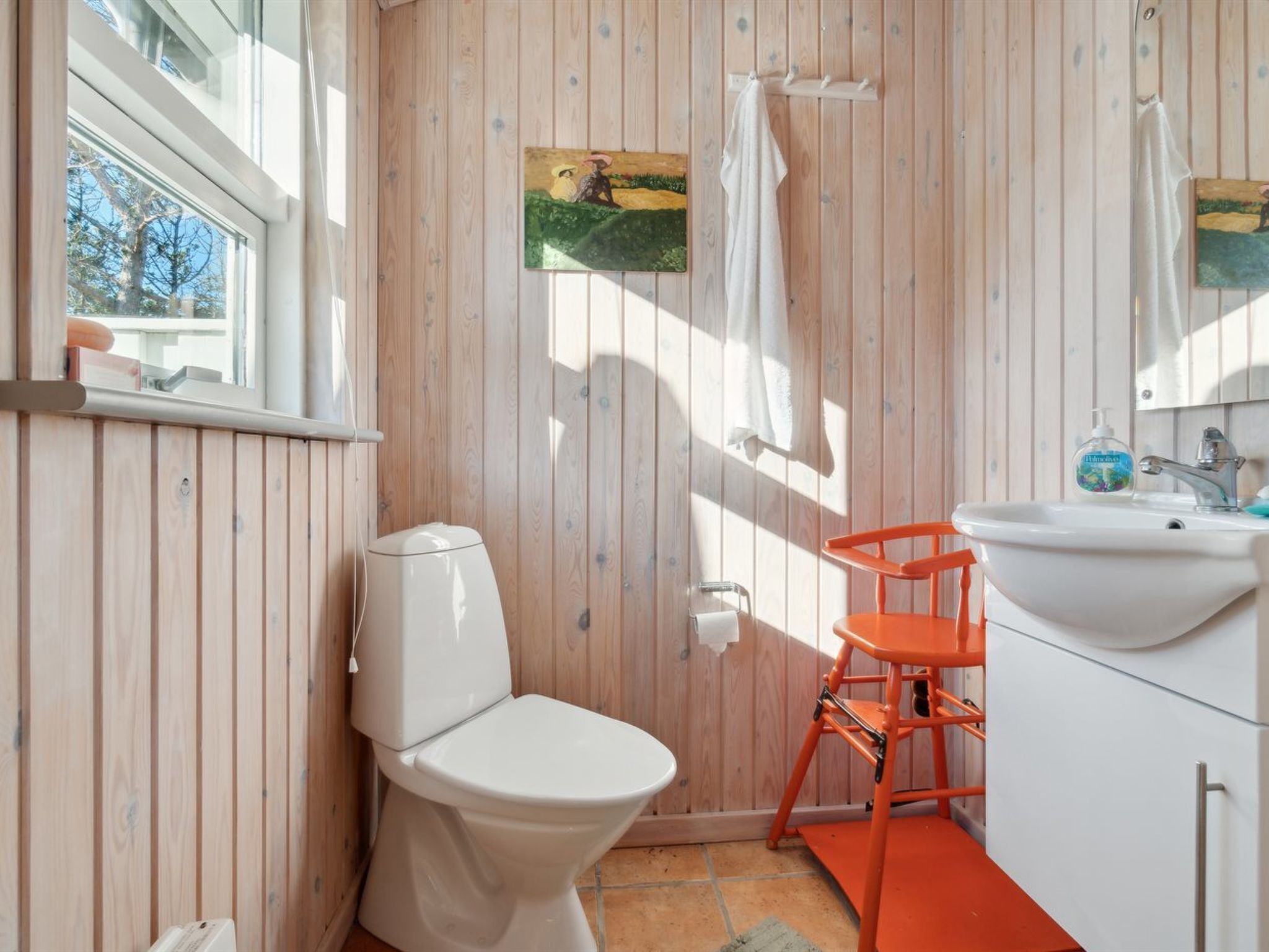 Ein Badezimmer mit Holzvertäfelung, einer weißen Toilette, einem orangefarbenen Stuhl, einem Waschbecken und einem Bild an der Wand.