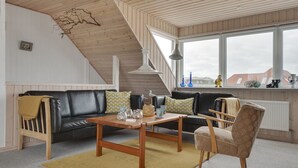Una sala de estar acogedora con un sofá de cuero negro, una mesa de centro de madera y un sillón estampado. Una escalera conduce a un nivel superior, y un ventanal ofrece vistas al exterior.