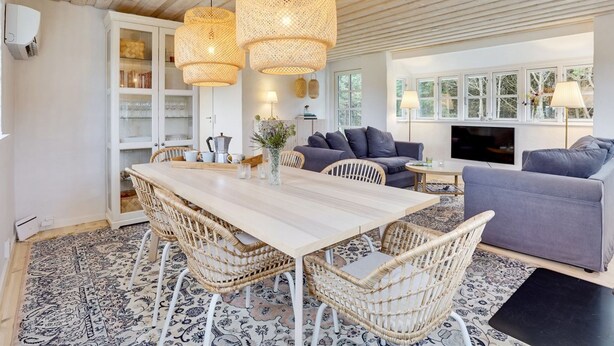 A spacious living area with a wooden ceiling, a dining table with wicker chairs, a grey sofa, and a TV.