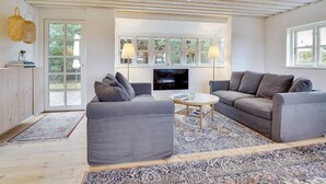 A modern living room with two grey sofas, a coffee table, and a TV. There are large windows with white frames and a wooden ceiling.