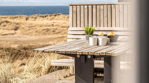A wooden deck with a table and chairs, overlooking a beach with sand dunes and the sea.