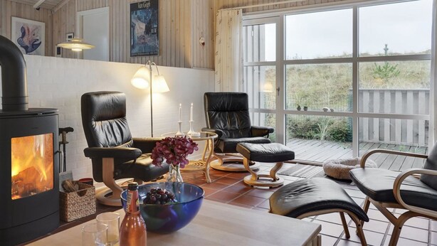 Un salon chaleureux avec un poêle à bois, des fauteuils en cuir, une table à manger ornée d’un panier de fruits et une grande fenêtre donnant sur un jardin.