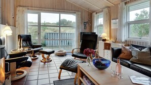 Une pièce chaleureuse avec un plafond en bois, de grandes fenêtres, un canapé, un fauteuil, une table avec un saladier et des verres, et une cheminée.