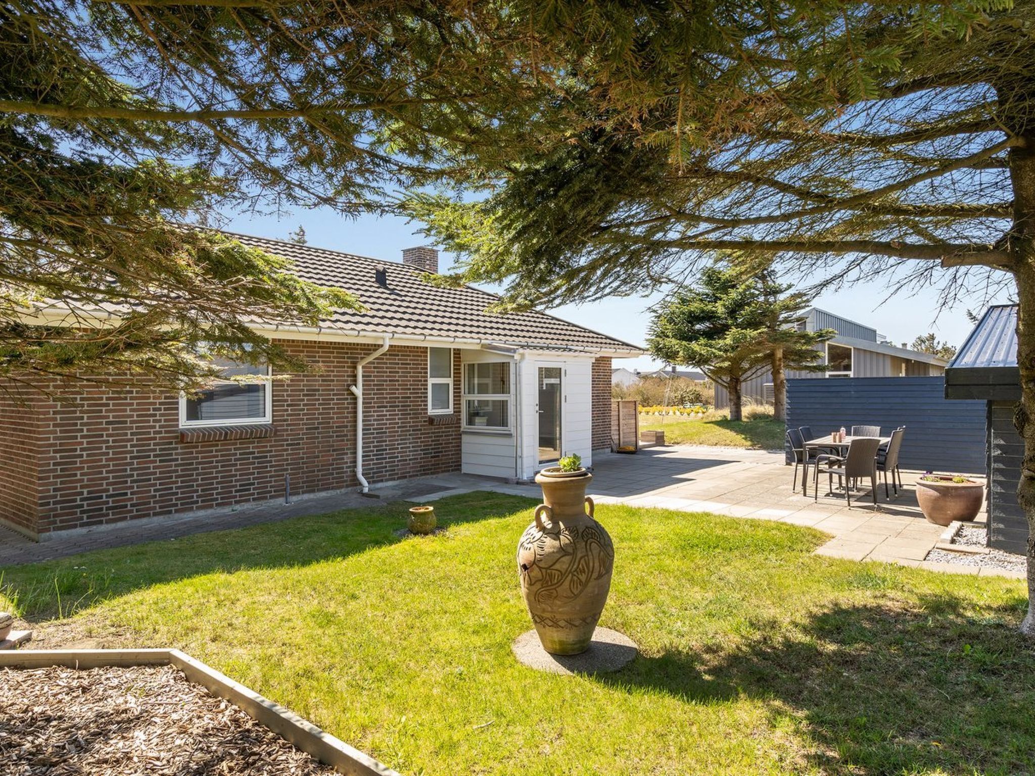 A residential backyard with a brick house, a patio with outdoor furniture, and a decorative vase.