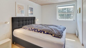 Een slaapkamer met een donker hoofdeinde, een bed met een decoratieve spreidek, een raam met uitzicht op bomen en een witte muur met ingelijste foto’s.