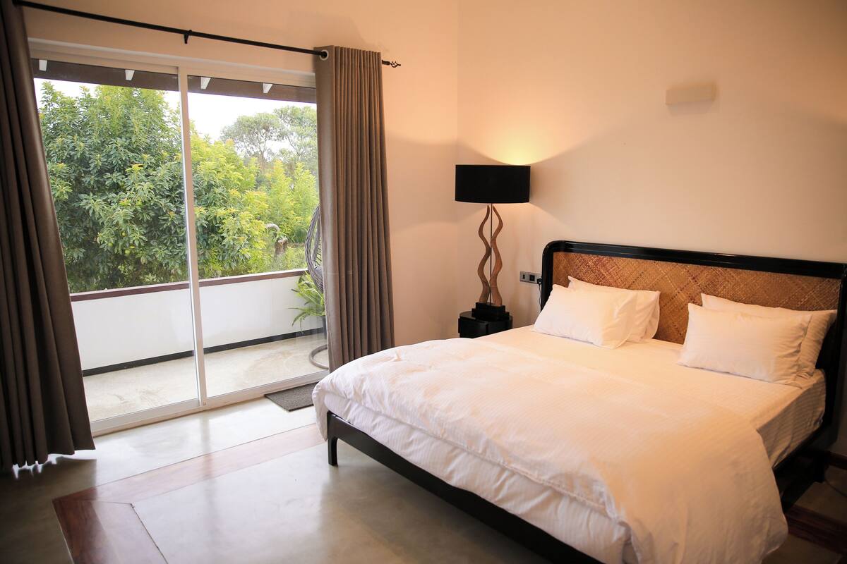 Quarto de hotel com uma cama grande, uma mesa de cabeceira com abajur e vista para o verde através de uma porta deslizante.