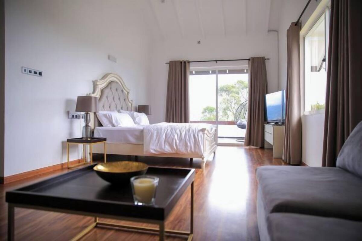 Quarto de hotel com cama, televisão, sofá, mesa de centro e janela com cortinas.