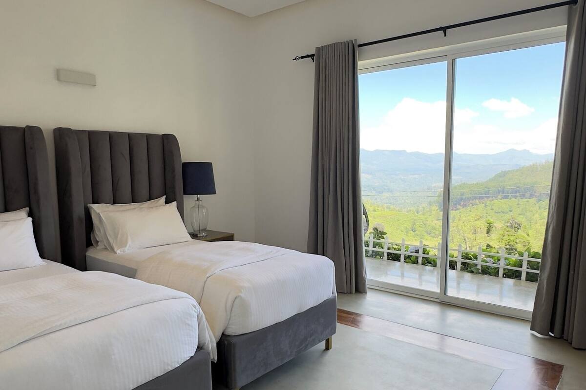 Quarto de hotel com duas camas, um ventilador de teto, uma janela grande com vista para montanhas e piso de madeira.