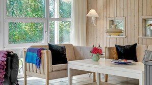 Un salon chaleureux avec un canapé, une table basse et une lampe. Il y a des fenêtres avec des rideaux, une cheminée et une vue sur la verdure à l’extérieur.