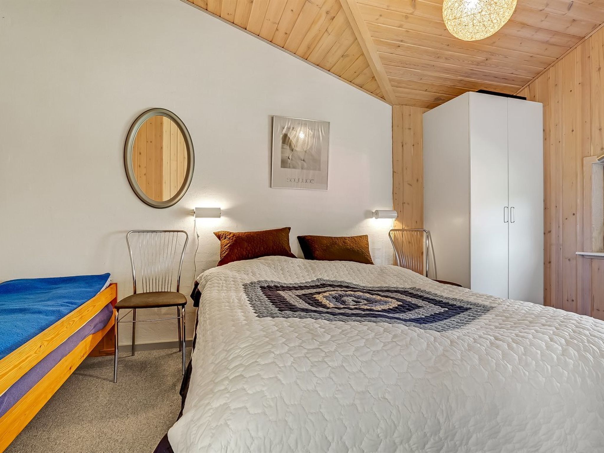 Een slaapkamer met een bed, een stoel, een kledingkast en een ronde spiegel aan de muur.