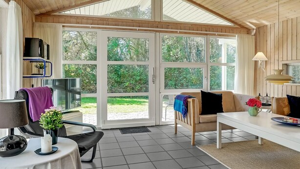 Une pièce spacieuse avec une grande porte-fenêtre coulissante donnant sur un jardin, une table blanche avec une lampe, un canapé et un fauteuil.