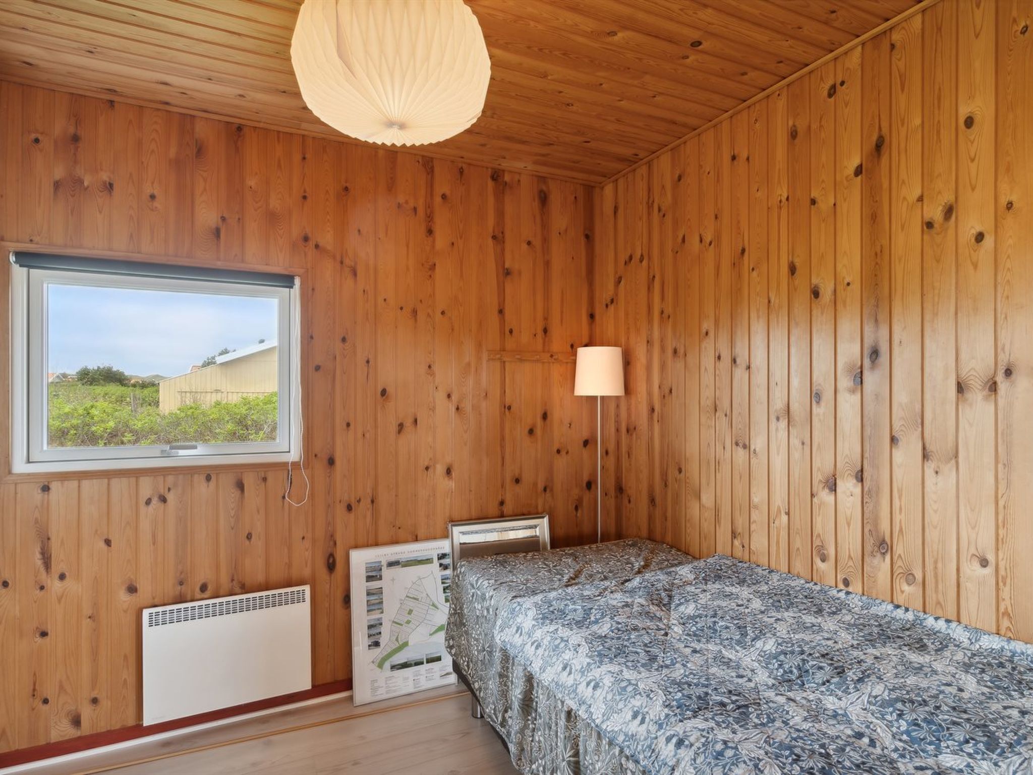 Une pièce aux murs en bois, une fenêtre donnant sur un beau paysage, un lit, une lampe sur pied et un radiateur.