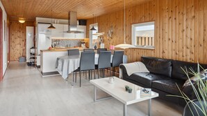 A modern interior with a wooden ceiling, a dining area with a white table and chairs, a black leather sofa, and a kitchen with wooden walls and a window.